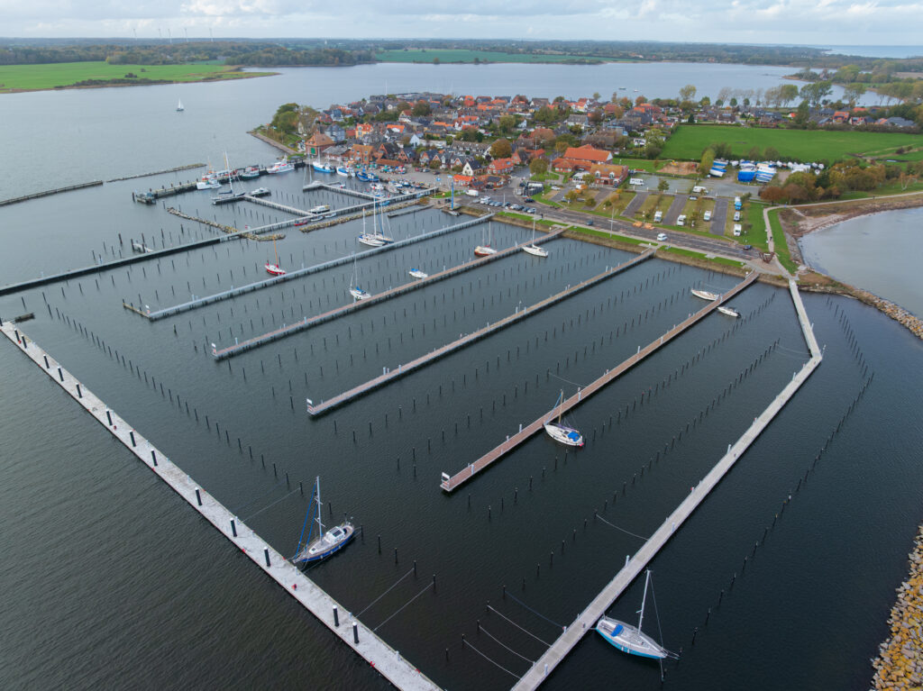 Luftaufnahme eines Jachthafens mit einigen angedockten Segelbooten und zahlreichen leeren Liegeplätzen, angrenzend an eine kleine Stadt und grüne Felder entlang der Küste.