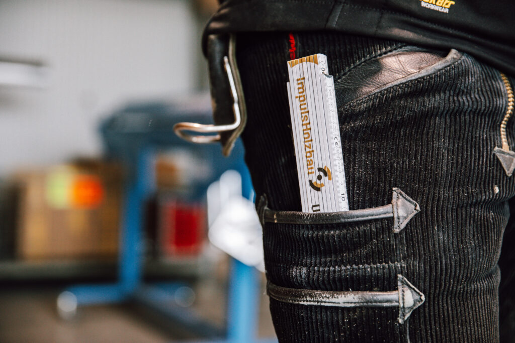 Nahaufnahme einer Person in schwarzer Arbeitshose mit einem Metalllineal mit der Aufschrift impulsoholzbau in einer Seitentasche; im Hintergrund ist eine unscharfe Werkstattumgebung zu sehen.