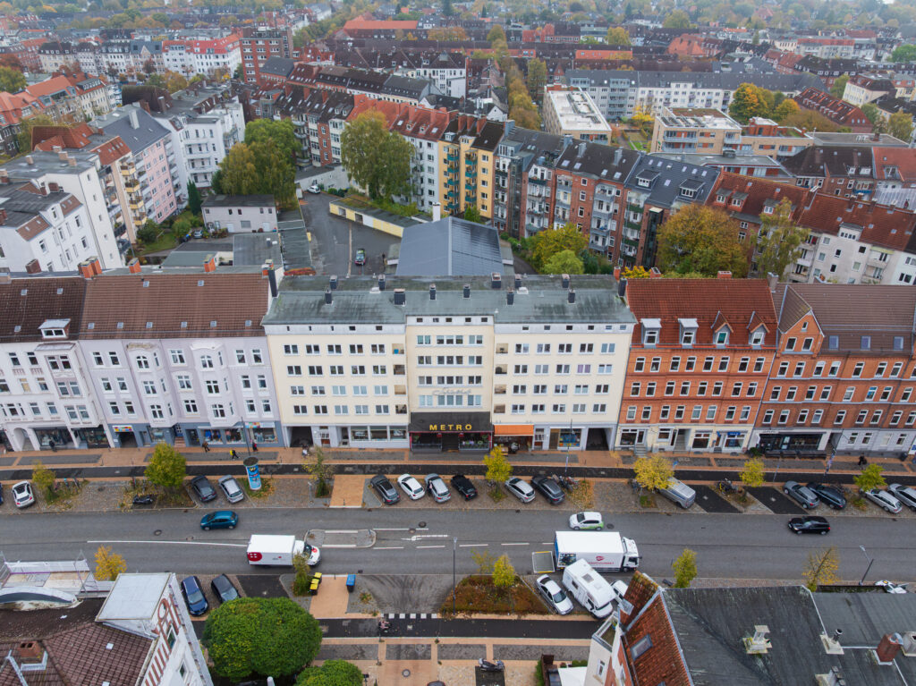 Luftaufnahme einer Stadtstraße mit geparkten Autos und mehrstöckigen Wohngebäuden mit roten und grauen Dächern. Im Hintergrund sind Bäume und weitere Gebäude zu sehen.