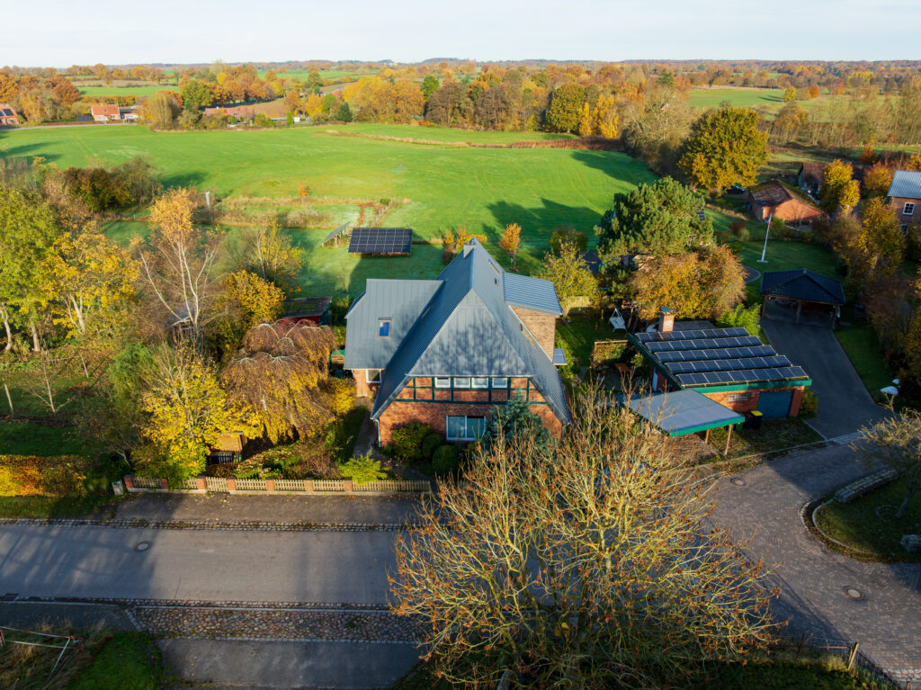 Luftaufnahme eines Hauses mit blauem Dach, umgeben von Bäumen und Feldern, mit Sonnenkollektoren auf nahe gelegenen Dächern und einer Straße im Vordergrund.