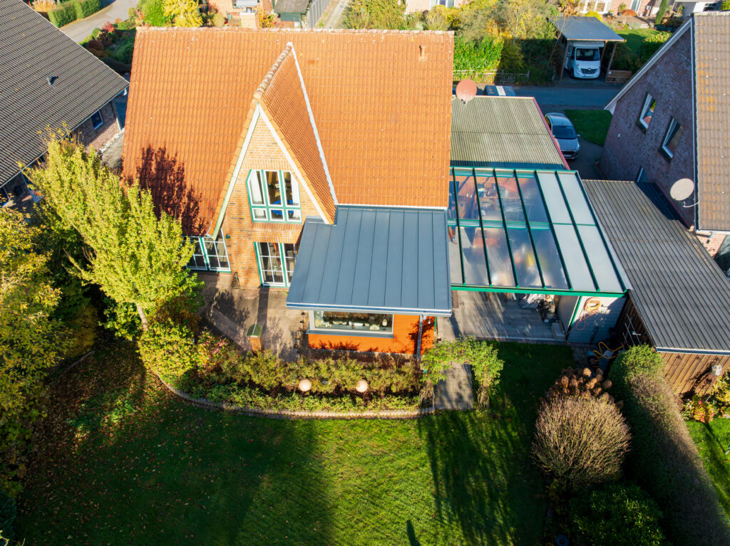 Luftaufnahme eines Hauses mit rotem Ziegeldach, glasüberdachter Terrasse, umliegendem Garten und angrenzender Einfahrt mit einem geparkten Auto.