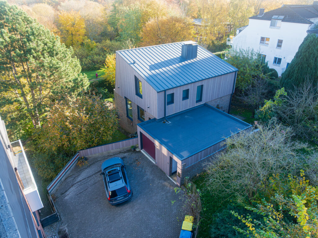 Luftaufnahme eines modernen zweistöckigen Hauses mit Flachdach und Holzfassade, einem in der Einfahrt geparkten Auto und umliegenden Bäumen mit Herbstlaub.